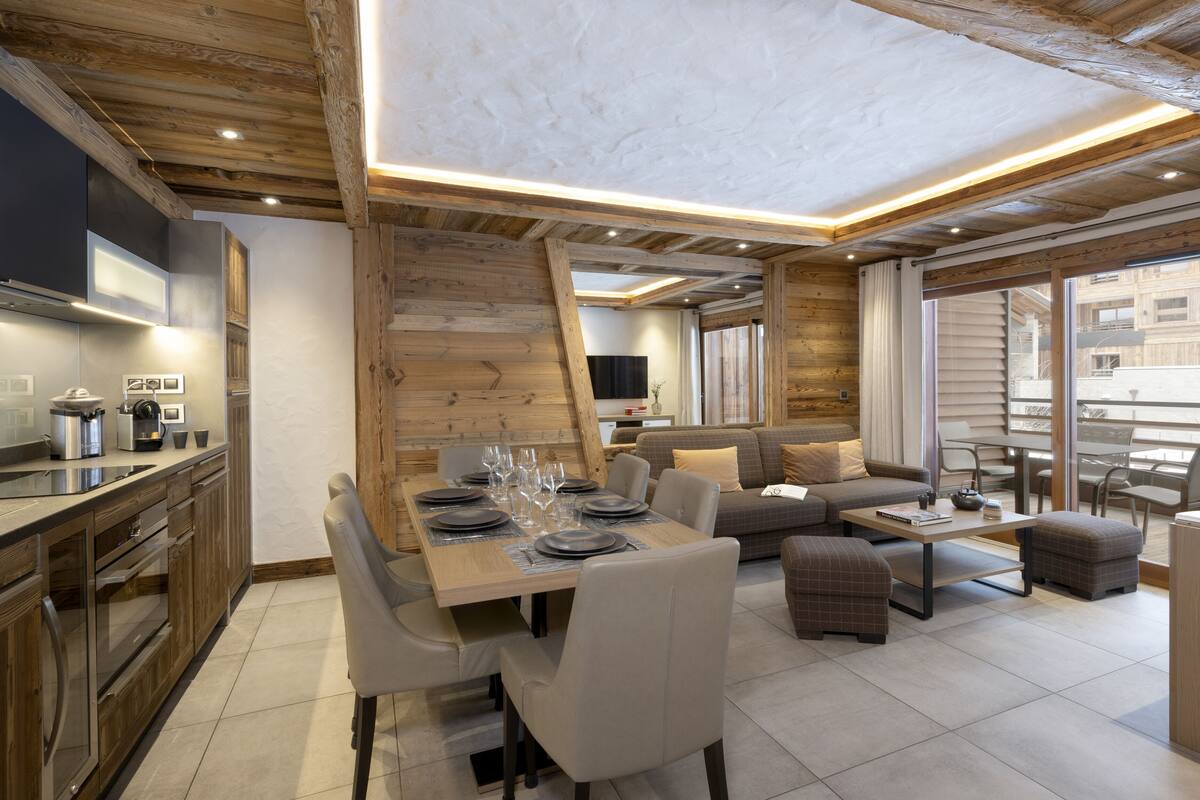 A modern kitchen with wooden cabinetry, a dining area with a table set for four, and a living area with a sofa and ottomans.