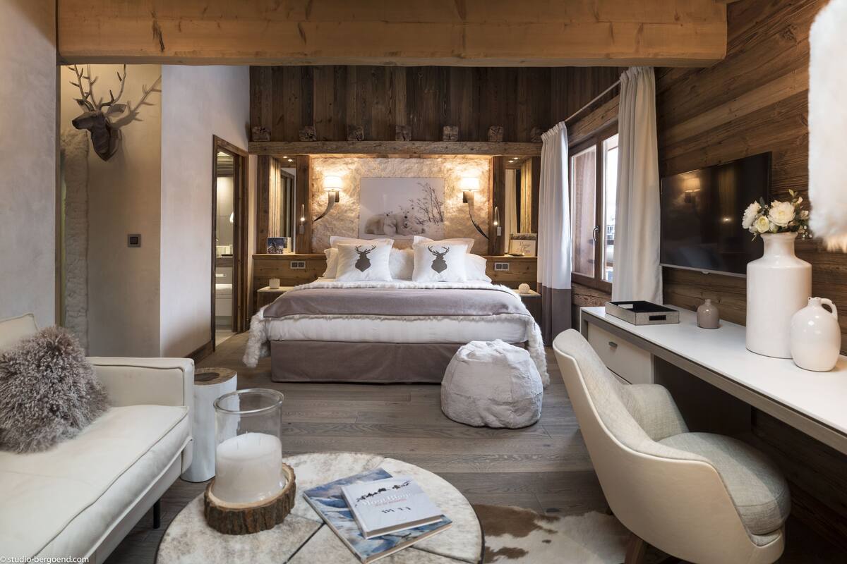 A cozy bedroom with a large bed, a desk, and a chair. There is a fireplace, a TV, and a window with curtains.