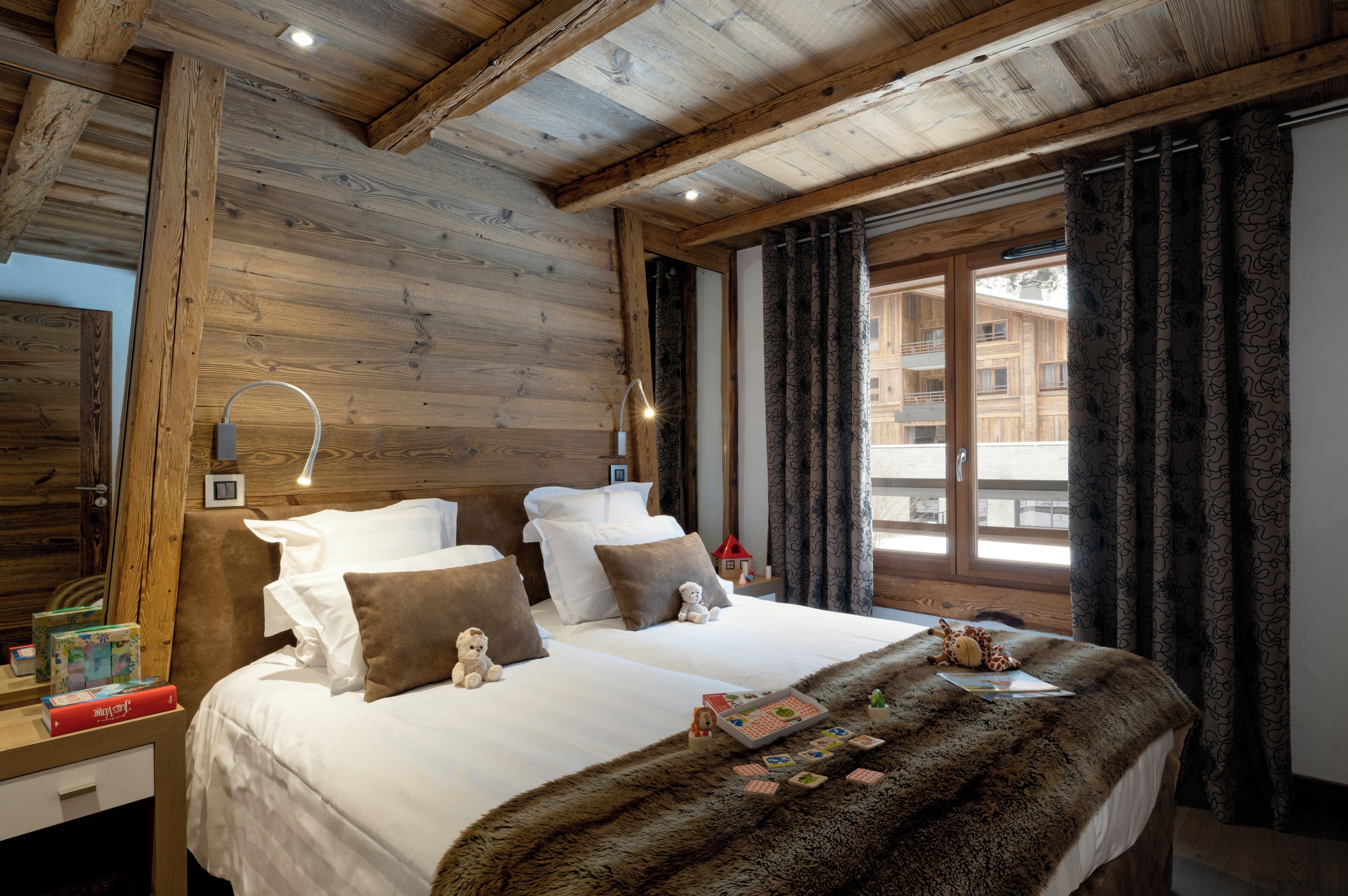 Une chambre confortable avec un plafond en bois, un grand lit recouvert de draps blancs et agrémenté d’un plaid en fourrure, une table de chevet avec des livres et une lampe, et une fenêtre ornée de rideaux à motifs.