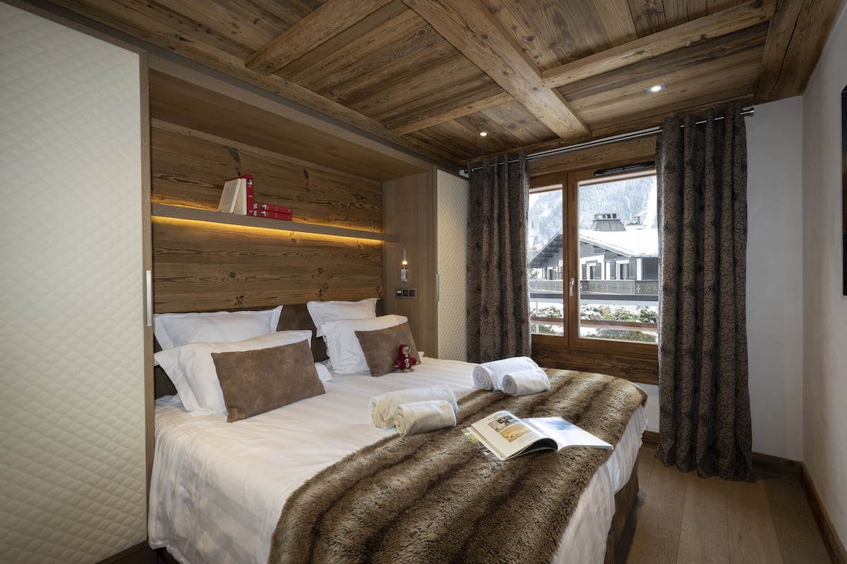 A bedroom with a wooden ceiling, a large bed with white and brown bedding, a fur throw blanket, and a window with patterned curtains.