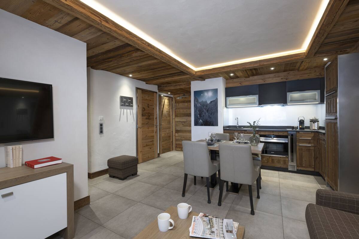 A modern kitchen with wooden cabinets, a dining area with a table and chairs, a flat-screen TV, and a cozy seating area with a sofa.