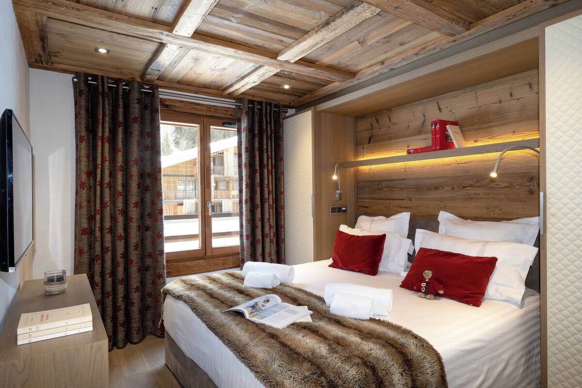 A bedroom with a wooden ceiling, a bed with white and red bedding, a bedside table with a book and a glass, and a window with curtains.