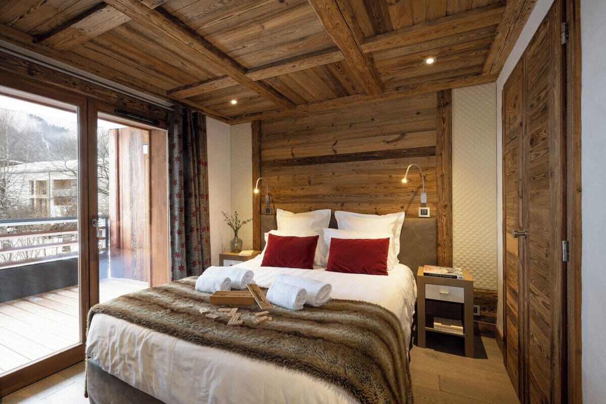 A wooden ceiling, a bed with red pillows, a nightstand, and a window with a view of a building.