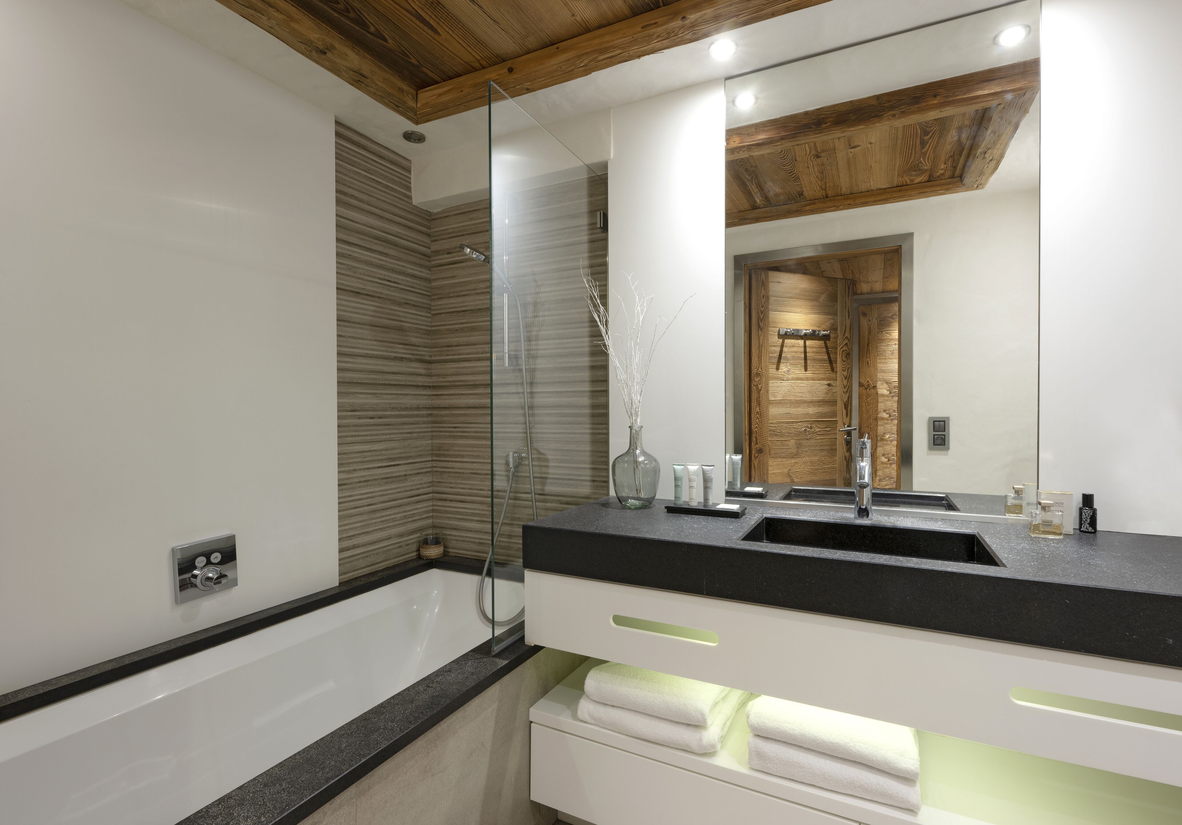 A modern bathroom with a glass shower enclosure, a wooden ceiling, and a large mirror reflecting a wooden door.
