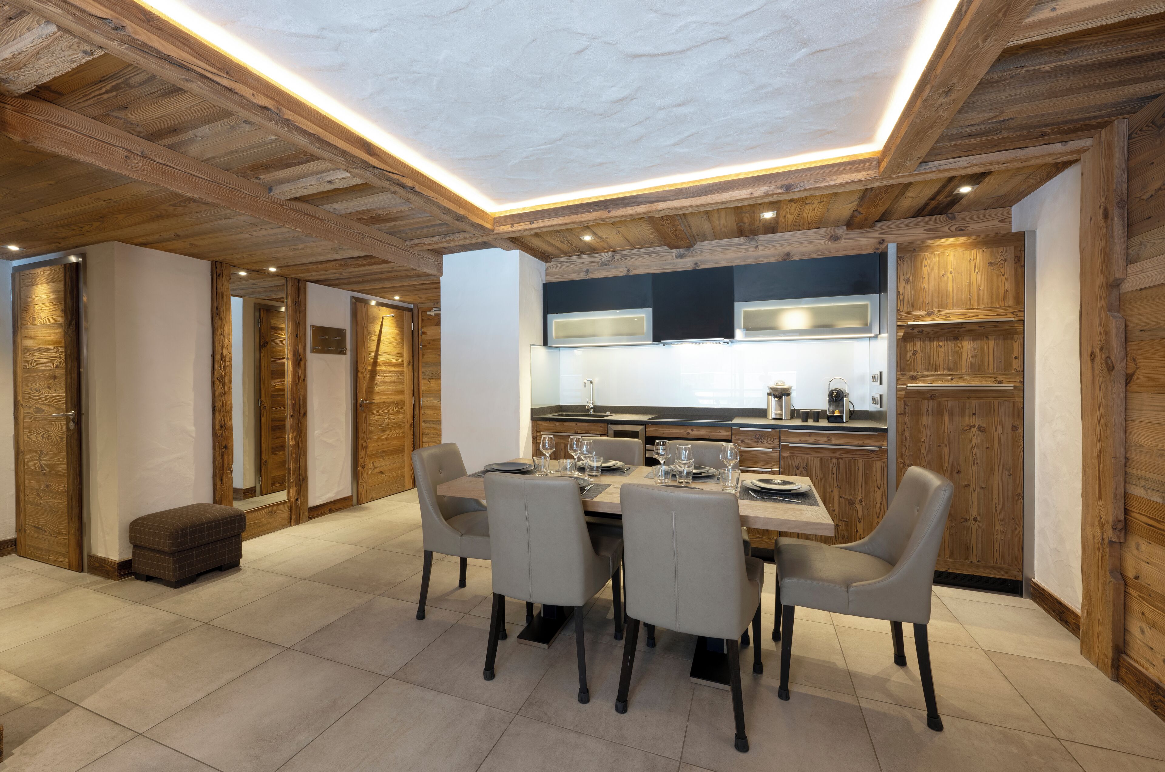A modern kitchen with wooden beams, a dining area with a table and chairs, and a small ottoman.