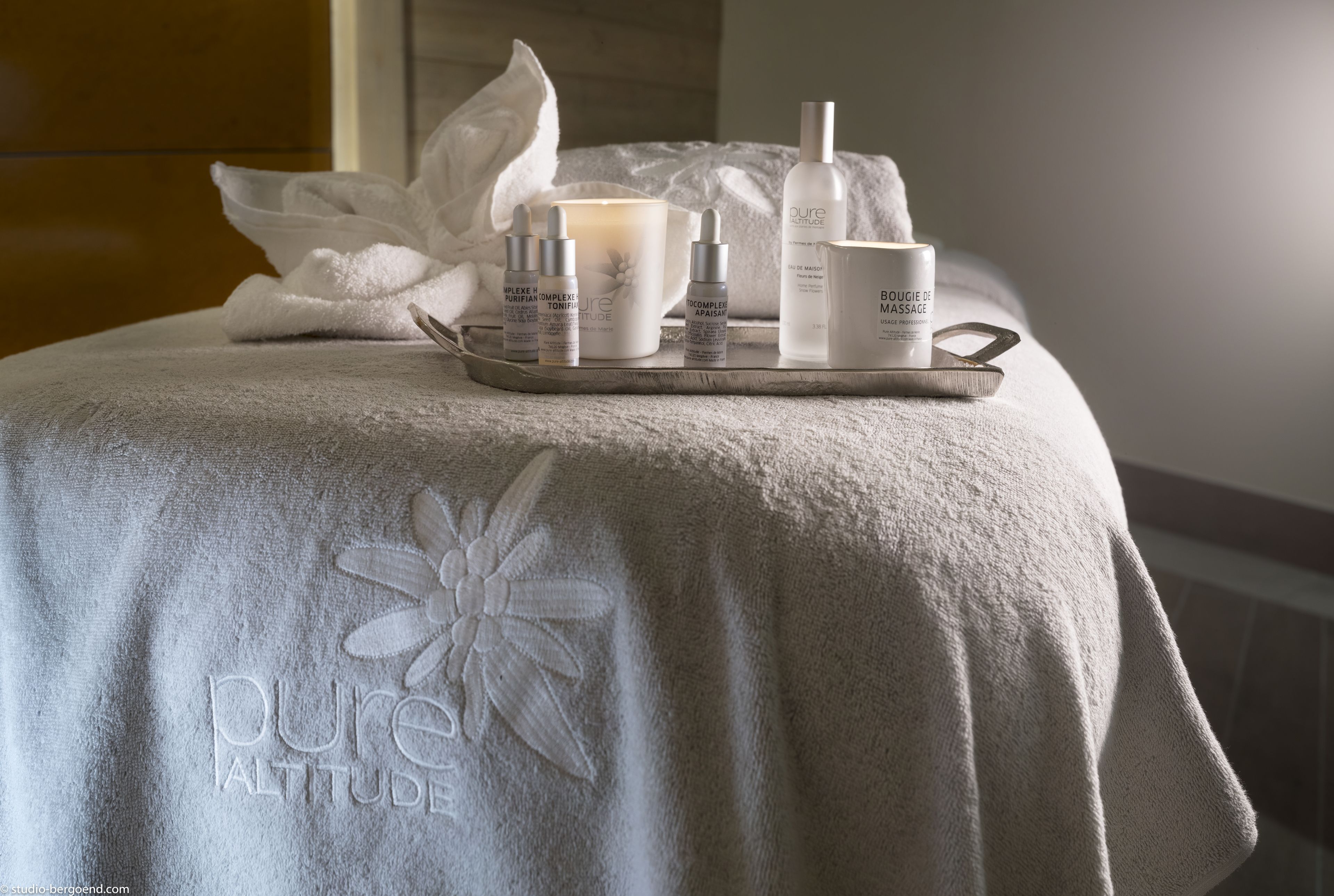 Un espace de bien-être avec une serviette ornée d’une broderie représentant le motif « pure altitude », un plateau contenant des produits de soins pour la peau, une bougie et une bouteille d’huile de massage « pure altitude ».