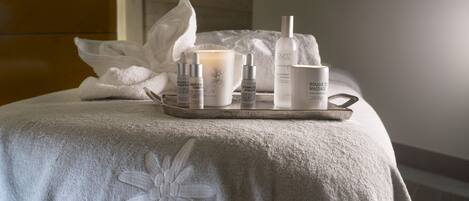 Un espace de bien-être avec une serviette ornée d’une broderie représentant le motif « pure altitude », un plateau contenant des produits de soins pour la peau, une bougie et une bouteille d’huile de massage « pure altitude ».