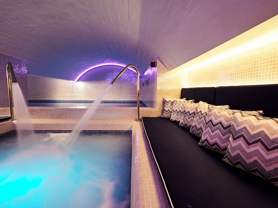 Um quarto de spa com hidromassagem, um sofá preto com almofadas estampadas e uma parede revestida com iluminação roxa.
