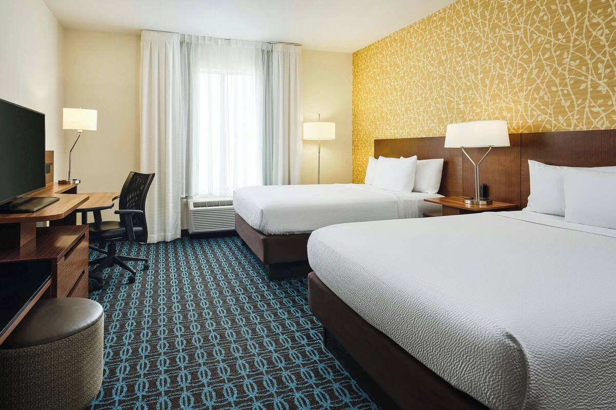 A hotel room with two beds, a desk with a chair, a TV, and a patterned carpet.