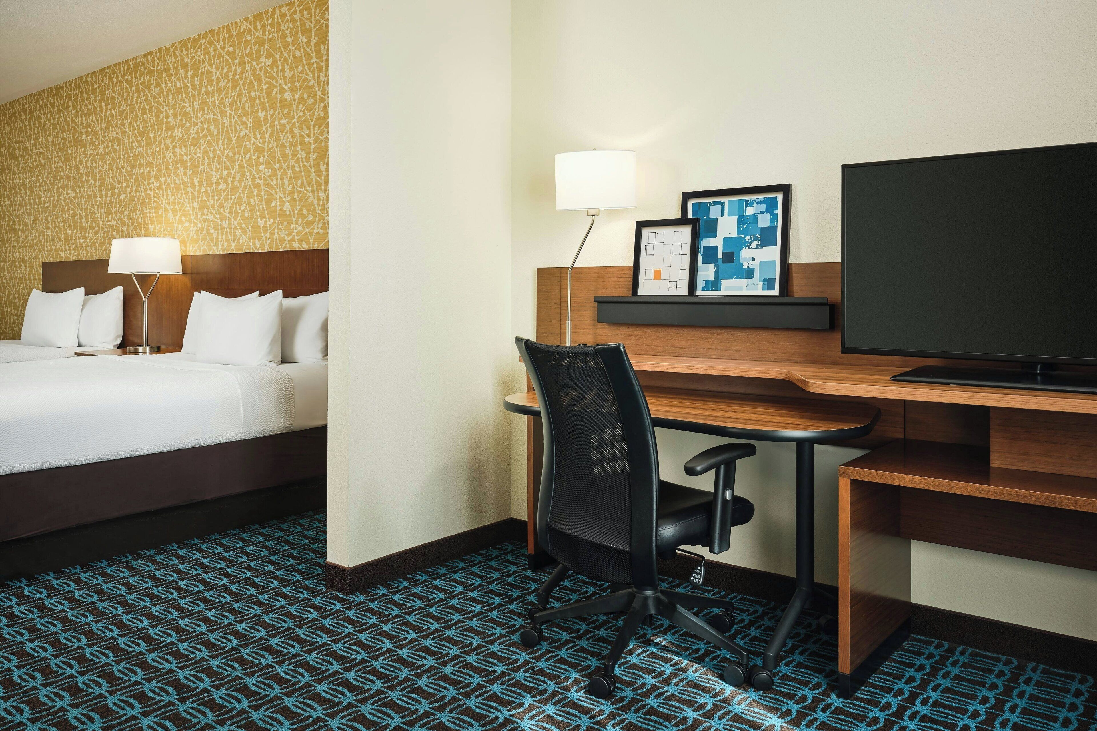 Habitación de hotel con cama, escritorio, silla, televisor, lámpara y una obra de arte enmarcada.
