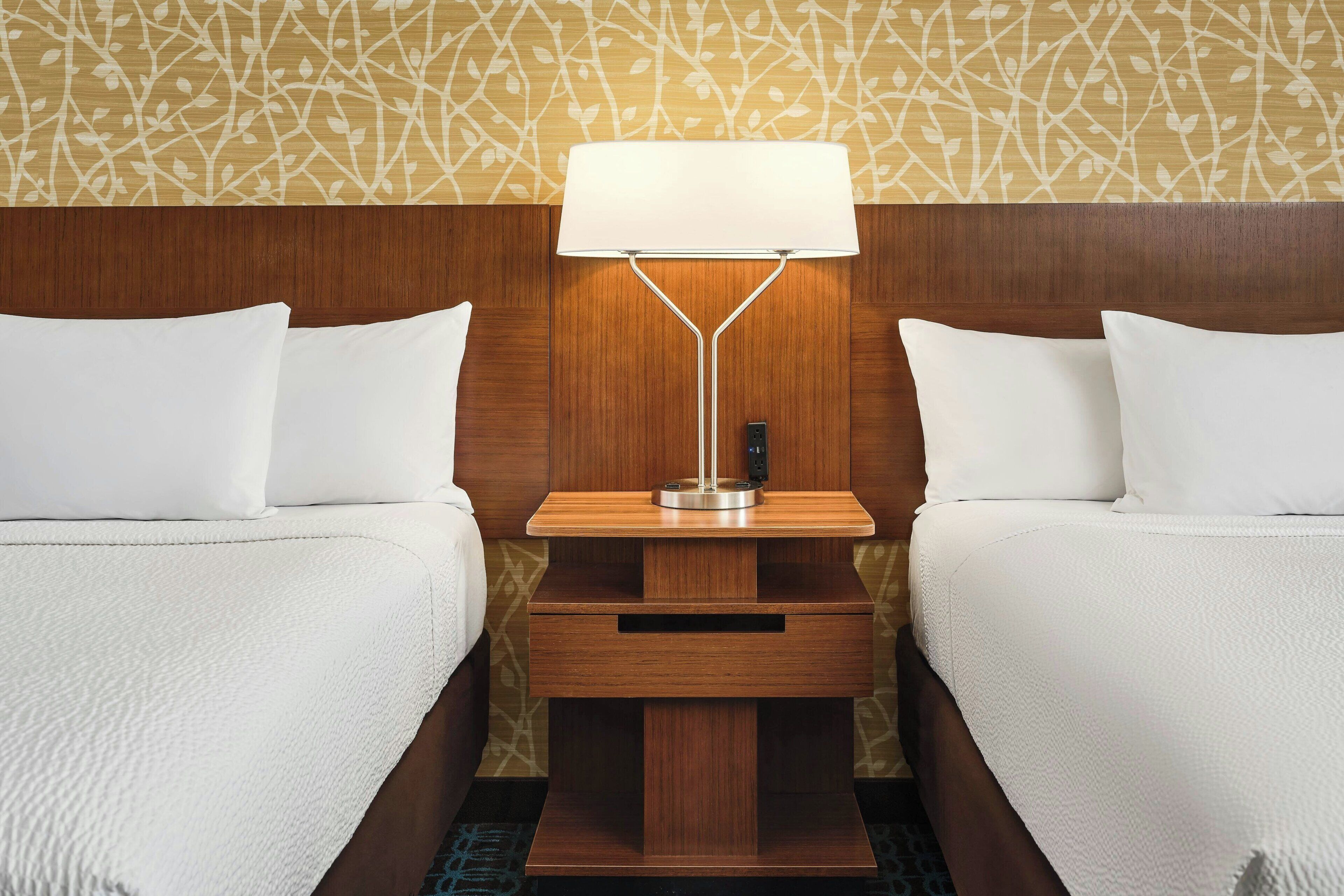 Habitación de hotel con dos camas, una mesita de noche con lámpara y pared con diseño.