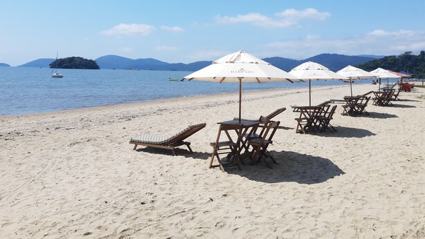 A beach with sun loungers and umbrellas, a boat in the water, and mountains in the background. - Casa Jardim Bangalôs (Paraty)