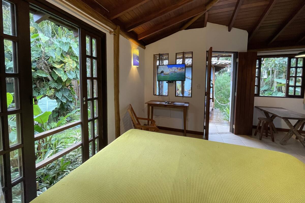 A room with a bed, a desk, and a TV. There are large windows and a view of greenery outside.