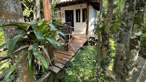 A wooden deck leading to a building with a tiled roof, surrounded by lush greenery and trees. - Casa Jardim Bangalôs (Paraty)
