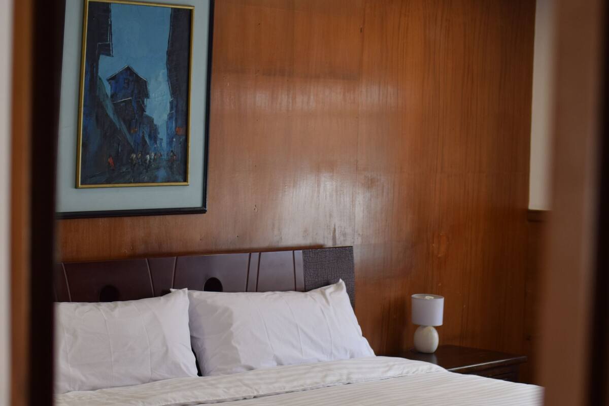 Un lit bien fait, avec des draps blancs, une tête de lit en bois et un tableau encadré accroché au mur.