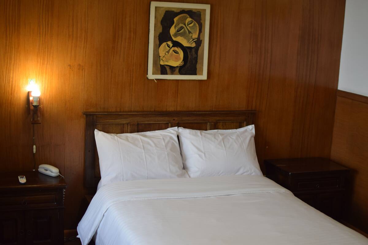 Une chambre d’hôtel avec une tête de lit en bois, un lit avec du linge de maison blanc, une table de chevet avec une lampe et un tableau au mur.