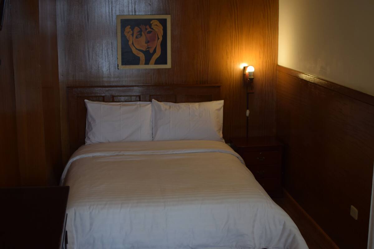 Un lit bien fait, avec du linge de lit blanc, dans une pièce aux murs lambrissés et dotée d’une seule table de chevet sur laquelle est posée une lampe fixée au mur.