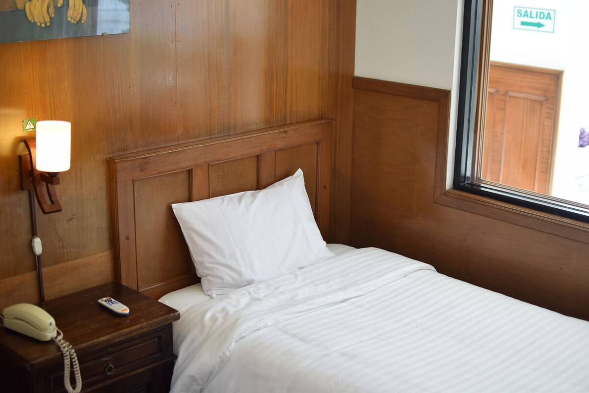 Une chambre d’hôtel avec une tête de lit en bois, un lit blanc, une lampe de chevet et un téléphone posé sur une table de chevet en bois.