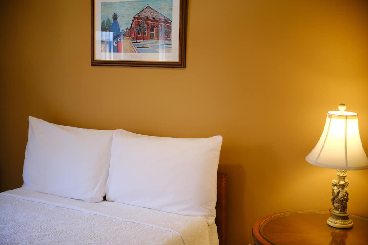 A neatly made bed with white linens, a framed painting above it, and a table lamp on a wooden side table.