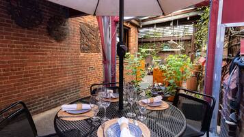 Una mesa metálica redonda dispuesta para comer, con sillas, una planta en maceta y un muro de ladrillo al fondo.