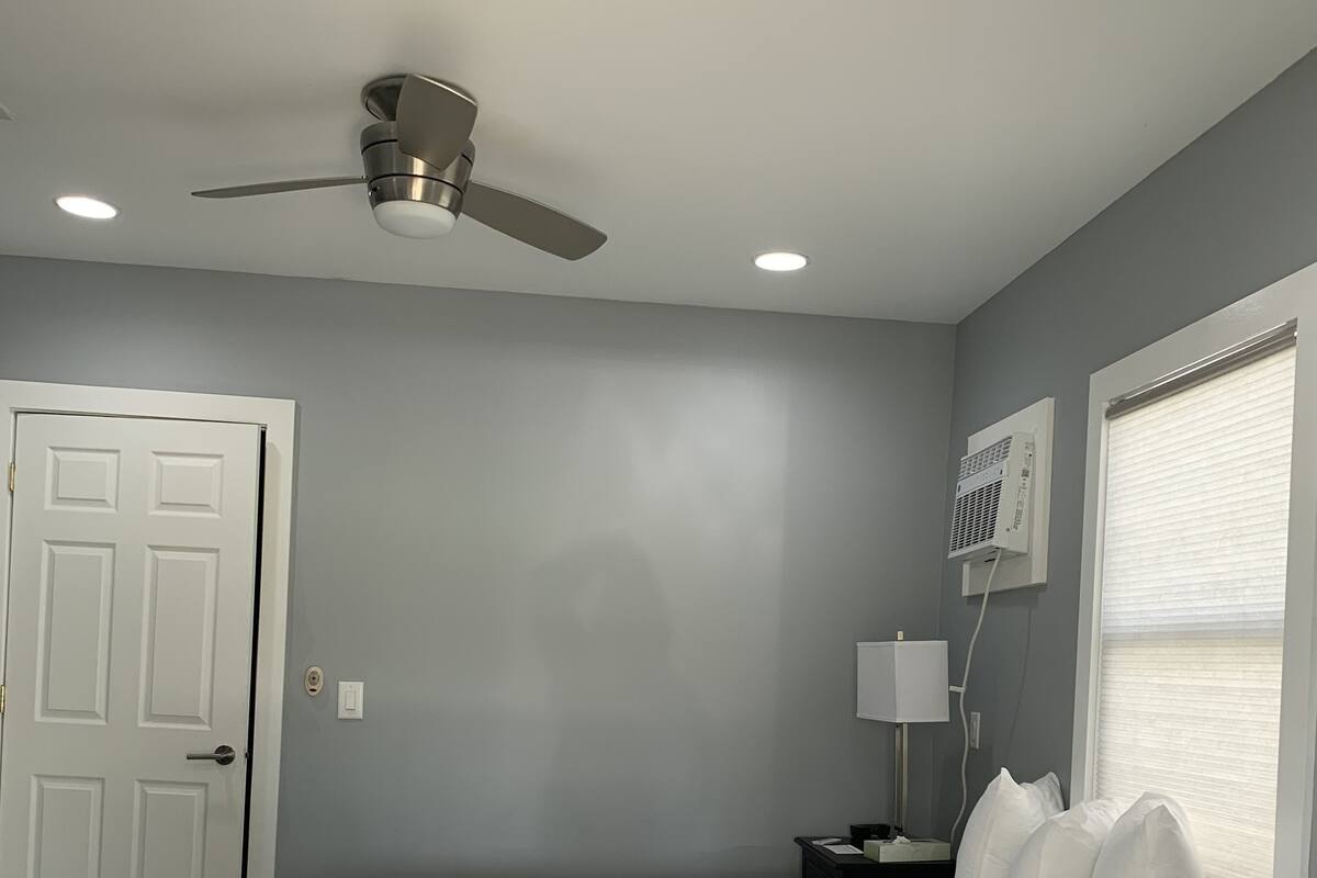 Een slaapkamer met een bed, een plafondventilator, inbouwverlichting, een deur, een nachtkastje met een lamp en een raam met jaloezieën.