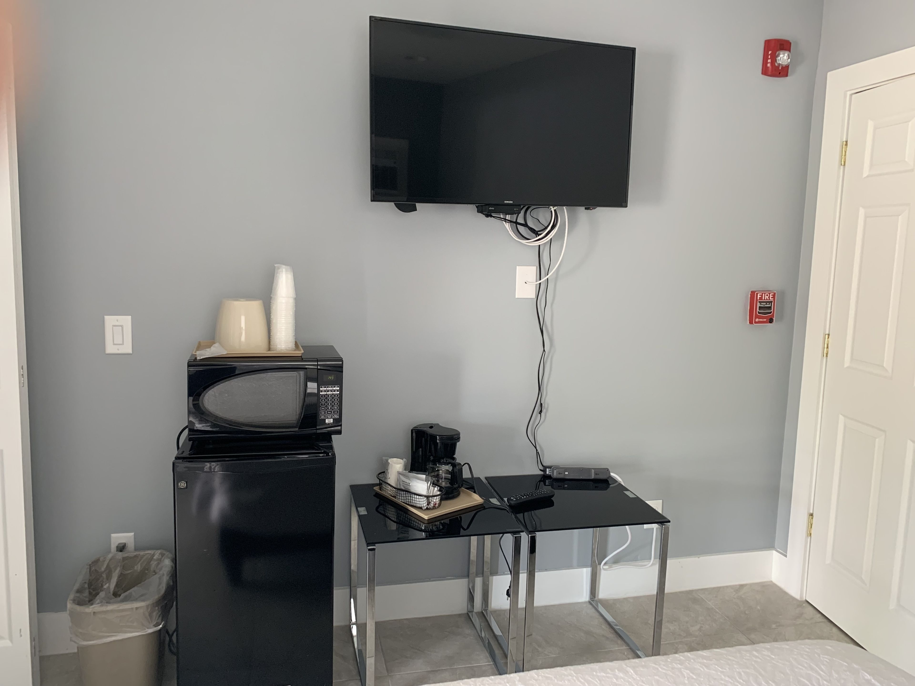 A compact room with a flat-screen TV mounted on the wall, a microwave, a small table with a coffee maker, and a trash bin.