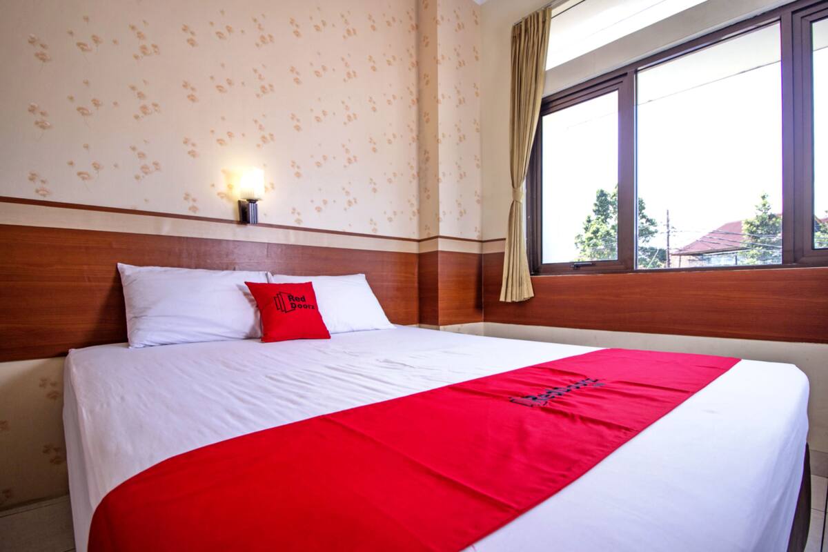 Une chambre d’hôtel avec un grand lit, un plaid rouge et une fenêtre donnant sur des immeubles.