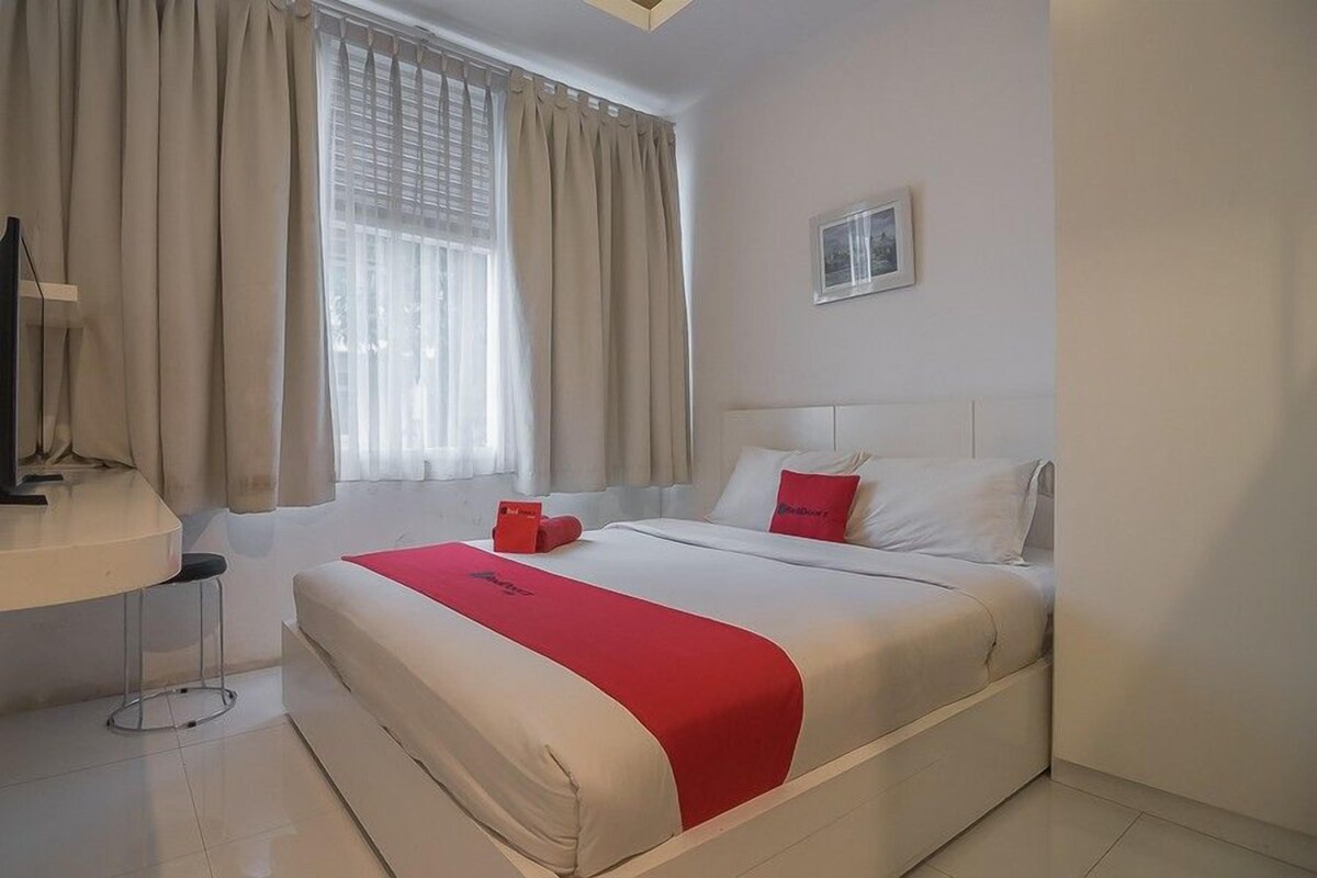 A hotel room with a bed, a bedside table, a chair, a window with curtains, and a picture on the wall.