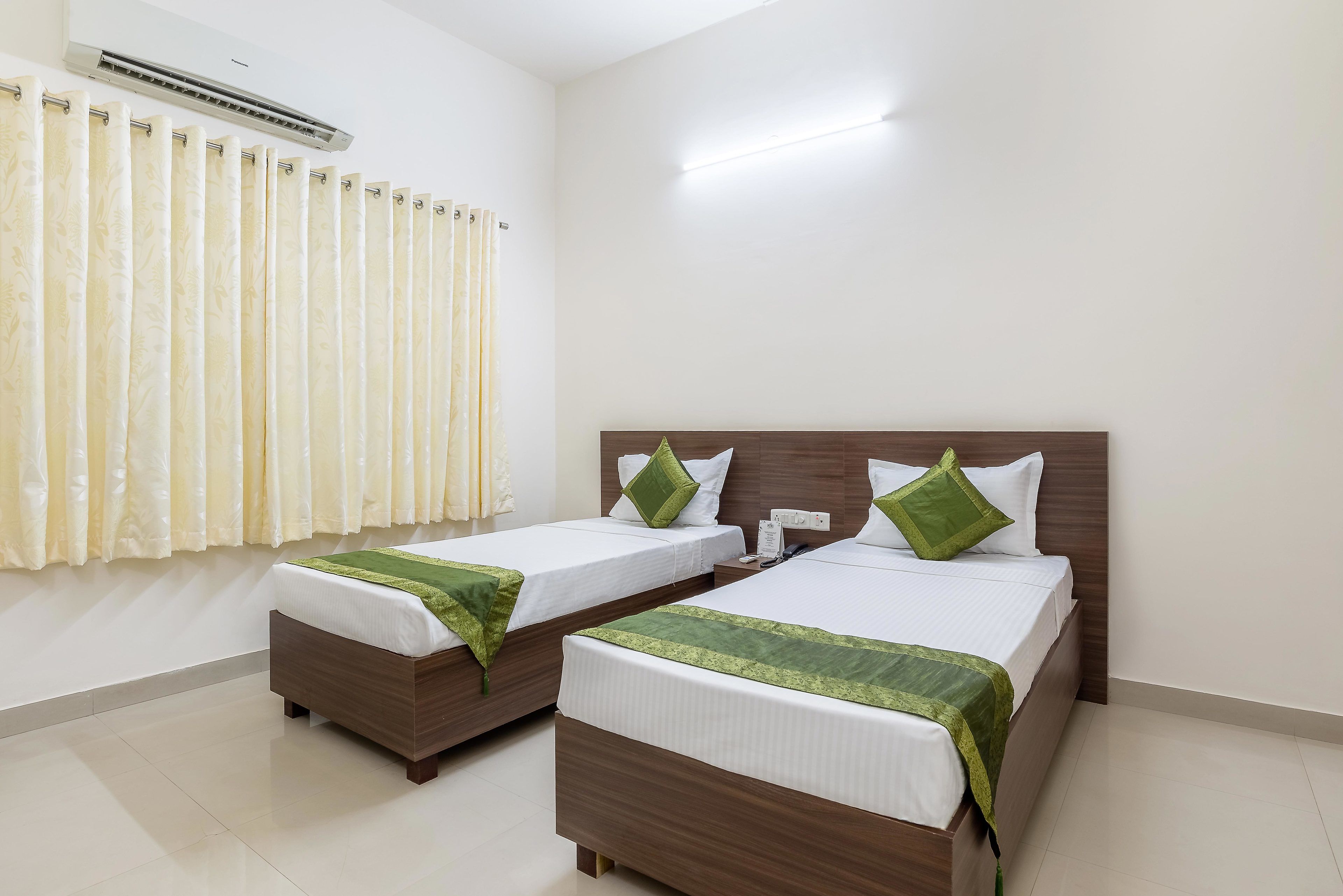 A hotel room with two single beds, a wooden headboard, a white wall, an air conditioning unit, and a curtain.