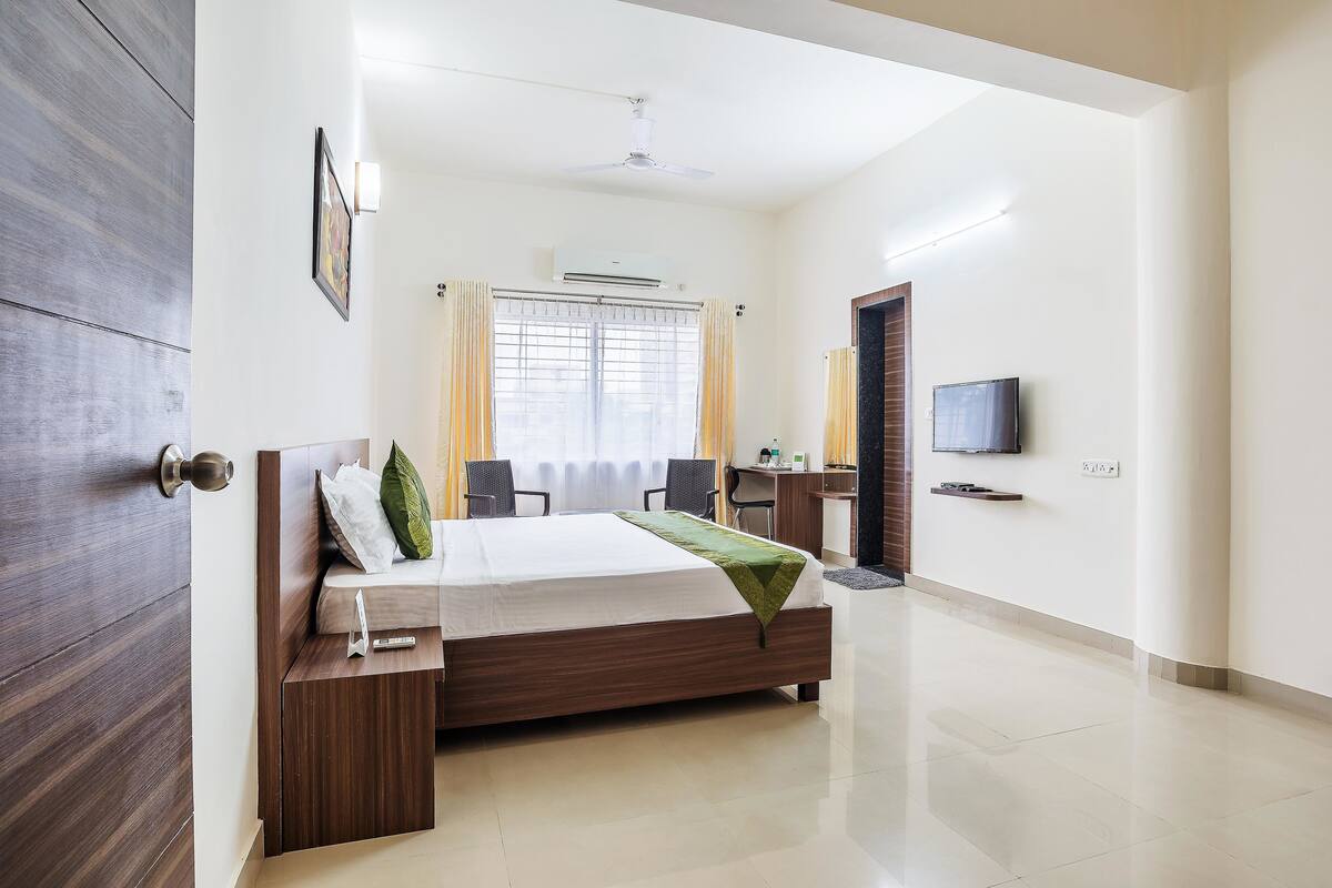 A hotel room with a bed, desk, chairs, a television, and a window with curtains.