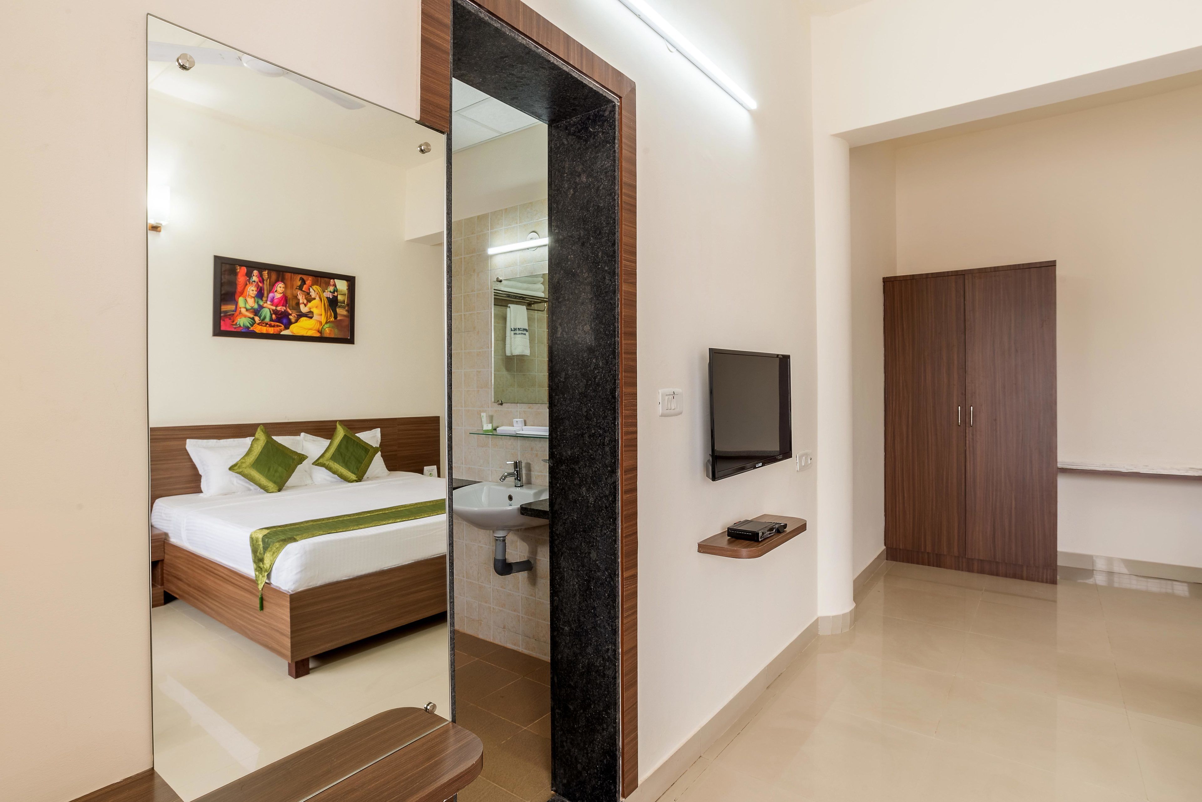 A hotel room with a bed, a mirror, a television, a wardrobe, and a bathroom visible through the mirror.