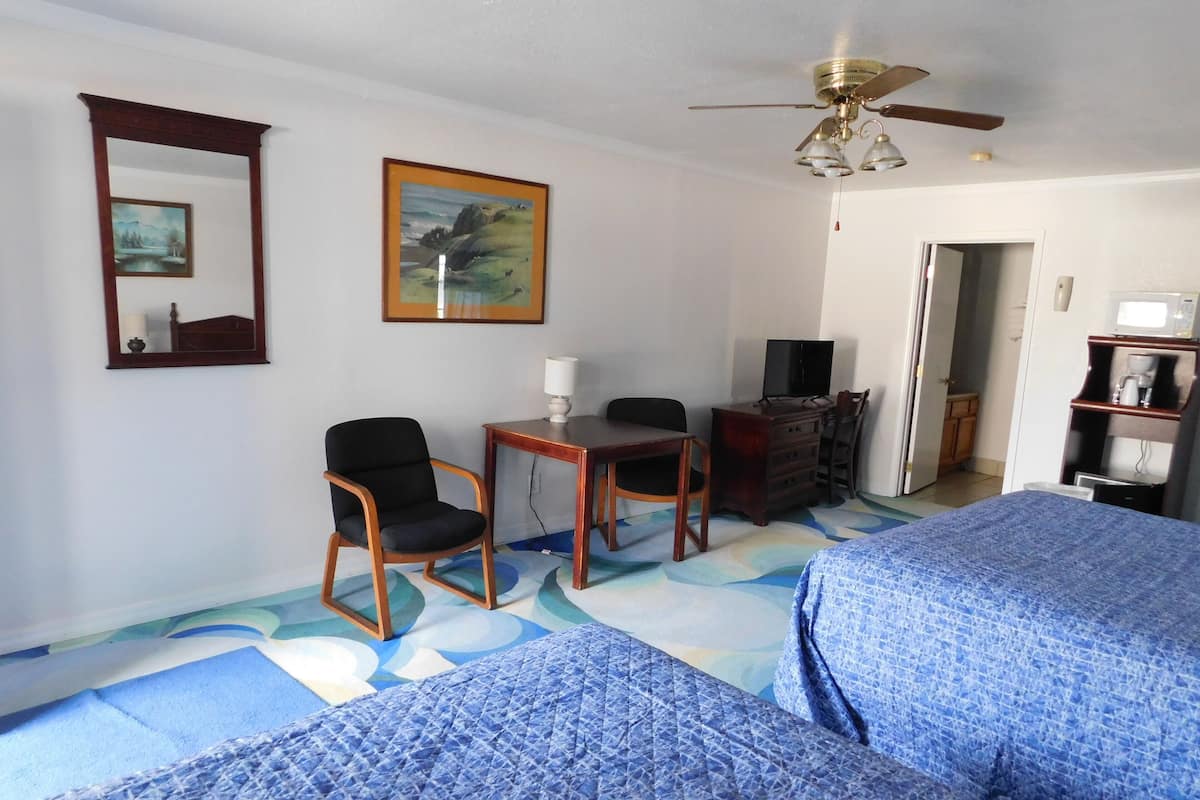 A hotel room with two beds, a desk with a chair, a television, a ceiling fan, and a painting on the wall.