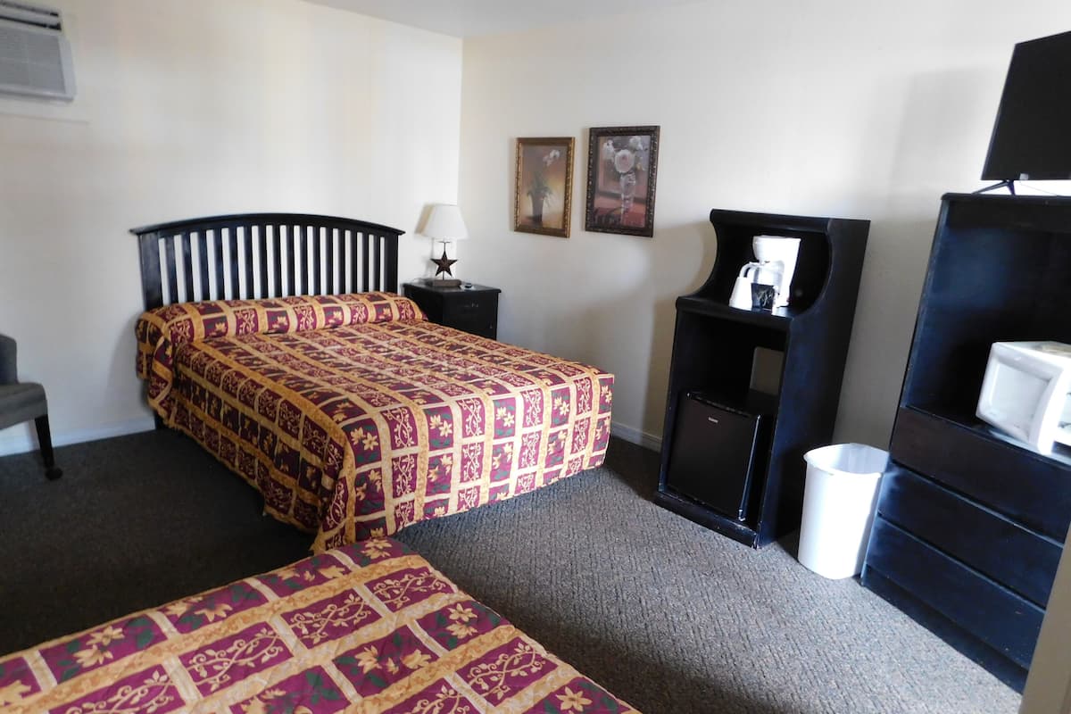 A hotel room with a bed, a small table with a lamp, a chair, a desk with a computer, and a bookshelf.