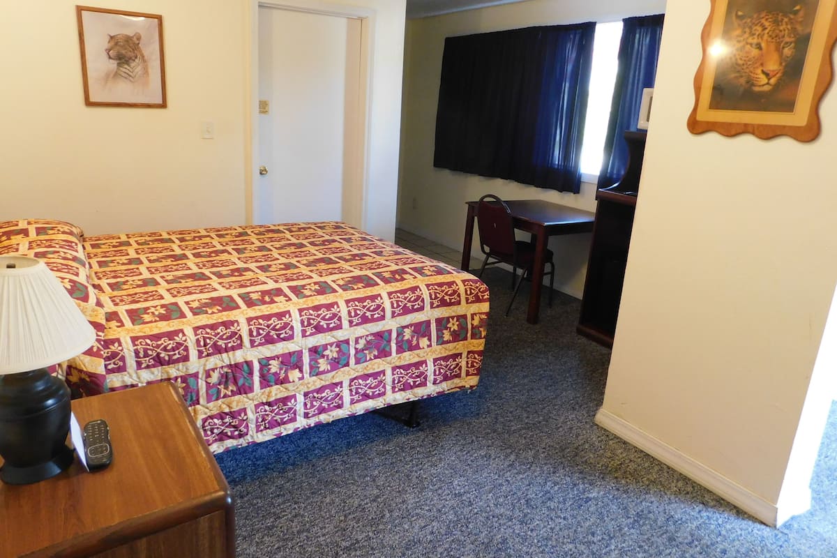 A hotel room with a bed, a desk, a chair, a lamp, and two framed pictures on the wall.
