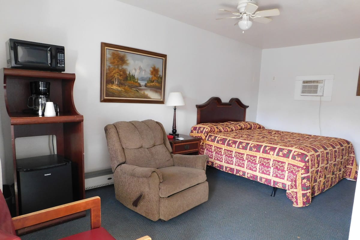 A hotel room with a bed, a chair, a desk with a computer, a coffee maker, a painting on the wall, and a ceiling fan.