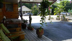 A wooden porch with outdoor seating, potted plants, and a view of a street with parked cars and a building in the background. - Little Shamrock Motel A1 (Murfreesboro)
