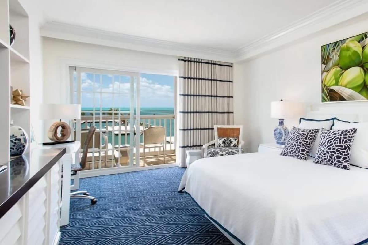 A hotel room with a bed, a chair, a desk, a lamp, and a view of the ocean.
