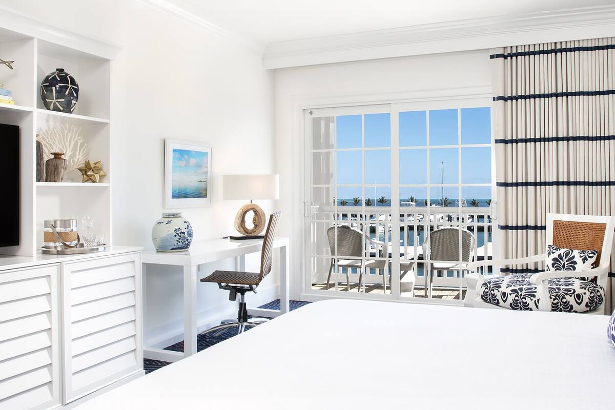 A hotel room with a bed, a desk with a chair, a window with a view of the ocean, and a balcony with chairs.