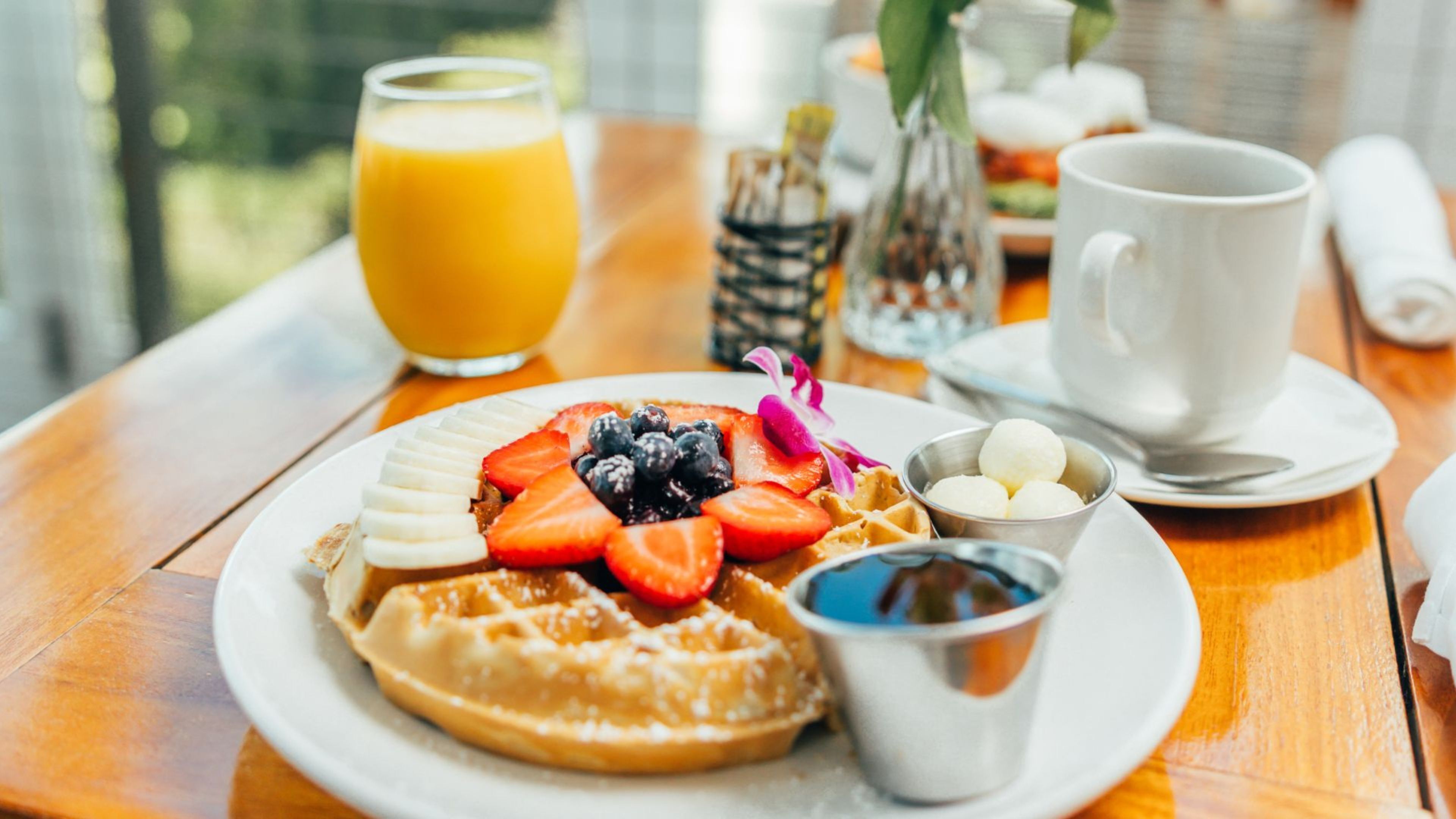 A breakfast plate with waffles, strawberries, blueberries, and a small bowl of syrup, accompanied by a glass of orange juice and a cup of coffee on a wooden table.