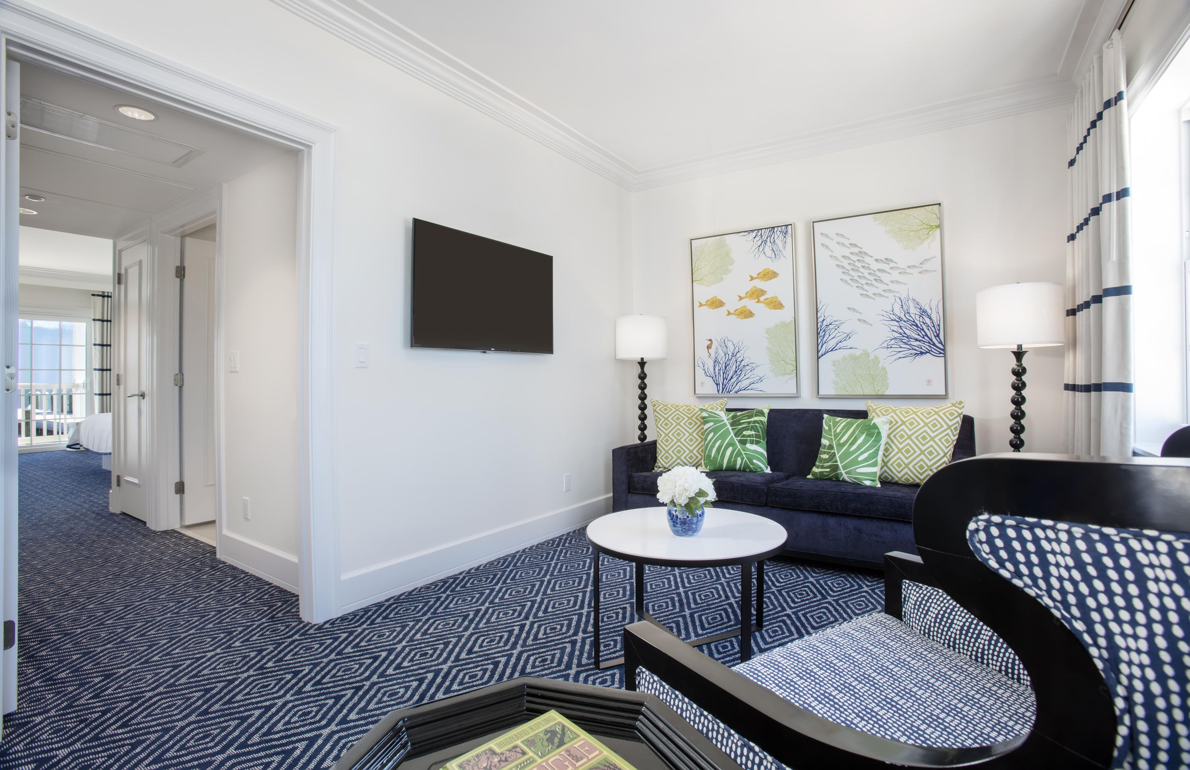 A hotel room with a flat-screen TV, two wall-mounted lamps, a sofa with patterned cushions, a round coffee table with a vase, and a patterned carpet.