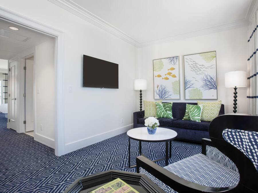 A hotel room with a flat-screen TV, two wall-mounted lamps, a sofa with patterned cushions, a round coffee table with a vase, and a patterned carpet.