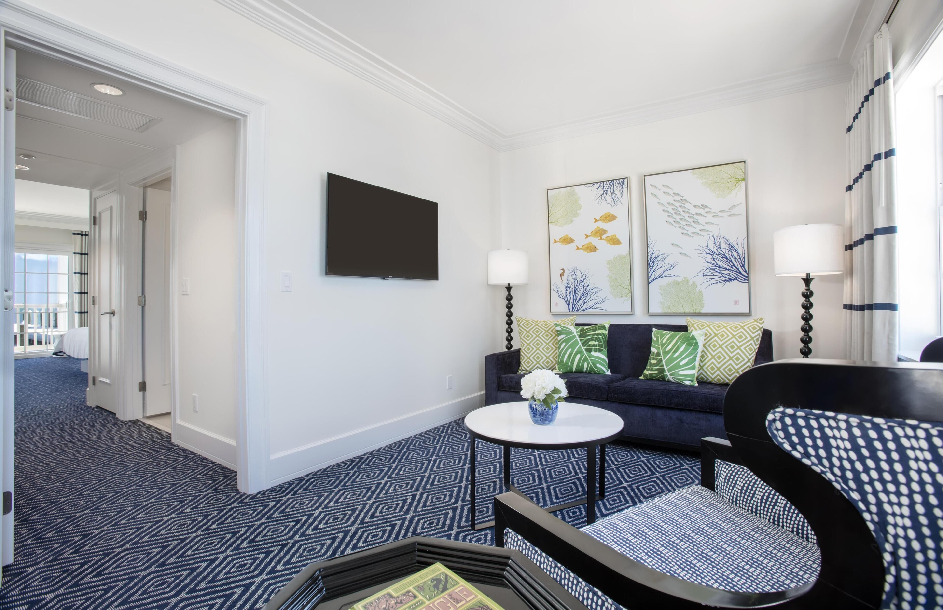 A hotel room with a flat-screen TV, two wall-mounted lamps, a sofa with patterned cushions, a round coffee table with a vase, and a patterned carpet.