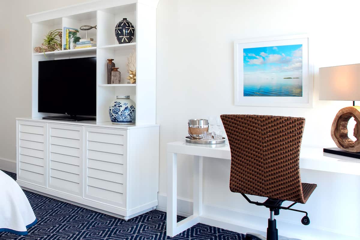 Une chambre d’hôtel avec un meuble de rangement blanc pour les appareils électroniques, un fauteuil en osier, un tableau encadré représentant une plage et une lampe.