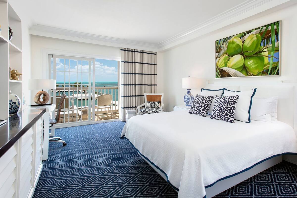 A hotel room with a large bed, a view of the ocean, a balcony with a chair, and a painting of coconuts.