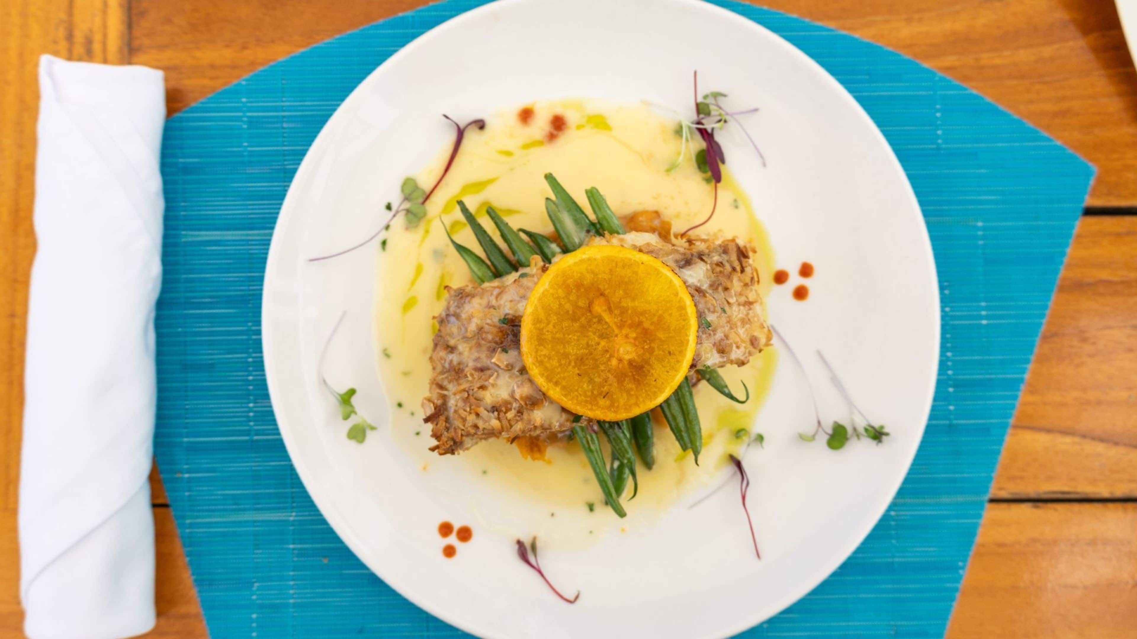 A white plate with a fish fillet, green garnish, and a slice of orange on a blue placemat.