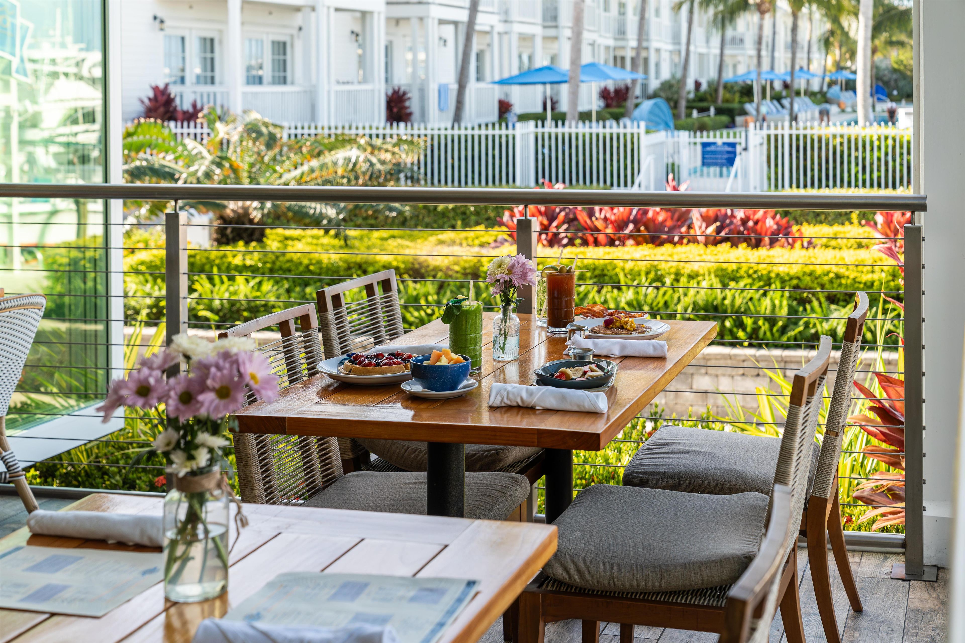 Un coin repas avec une table dressée pour deux personnes, des chaises, un vase de fleurs et une vue sur un jardin et des bâtiments.