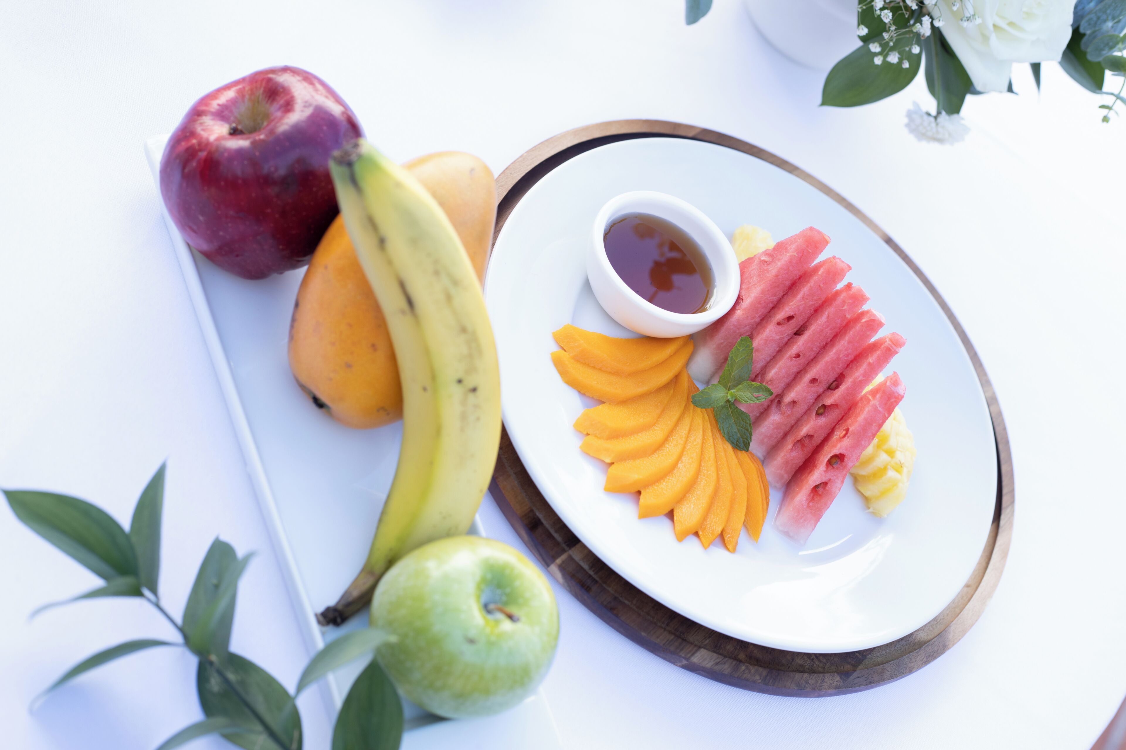 A plate with sliced fruits and a small bowl of syrup, accompanied by whole fruits and a decorative arrangement.
