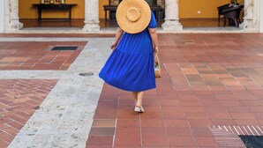 A woman in a blue dress and straw hat walking towards a building with a brick facade, arched doorways, and a balcony with flowers.