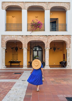 A woman in a blue dress and straw hat walking towards a building with a brick facade, arched doorways, and a balcony with flowers.