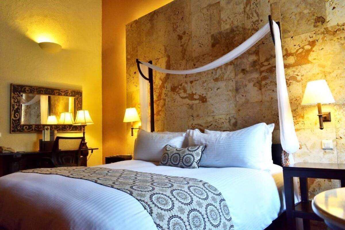 A hotel room with a four-poster bed, a desk with a chair, two wall-mounted lamps, and a mirror.